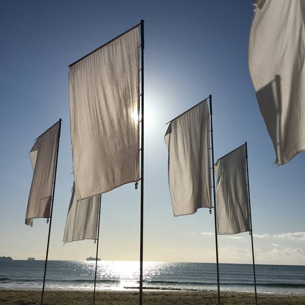 Banderas blancas en mástiles a contraluz, con el sol y el mar al fondo