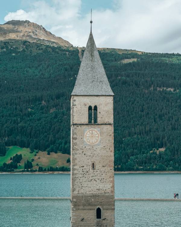 Torre de iglesia que emerge de un lago, con un bosque y una montaña detrás