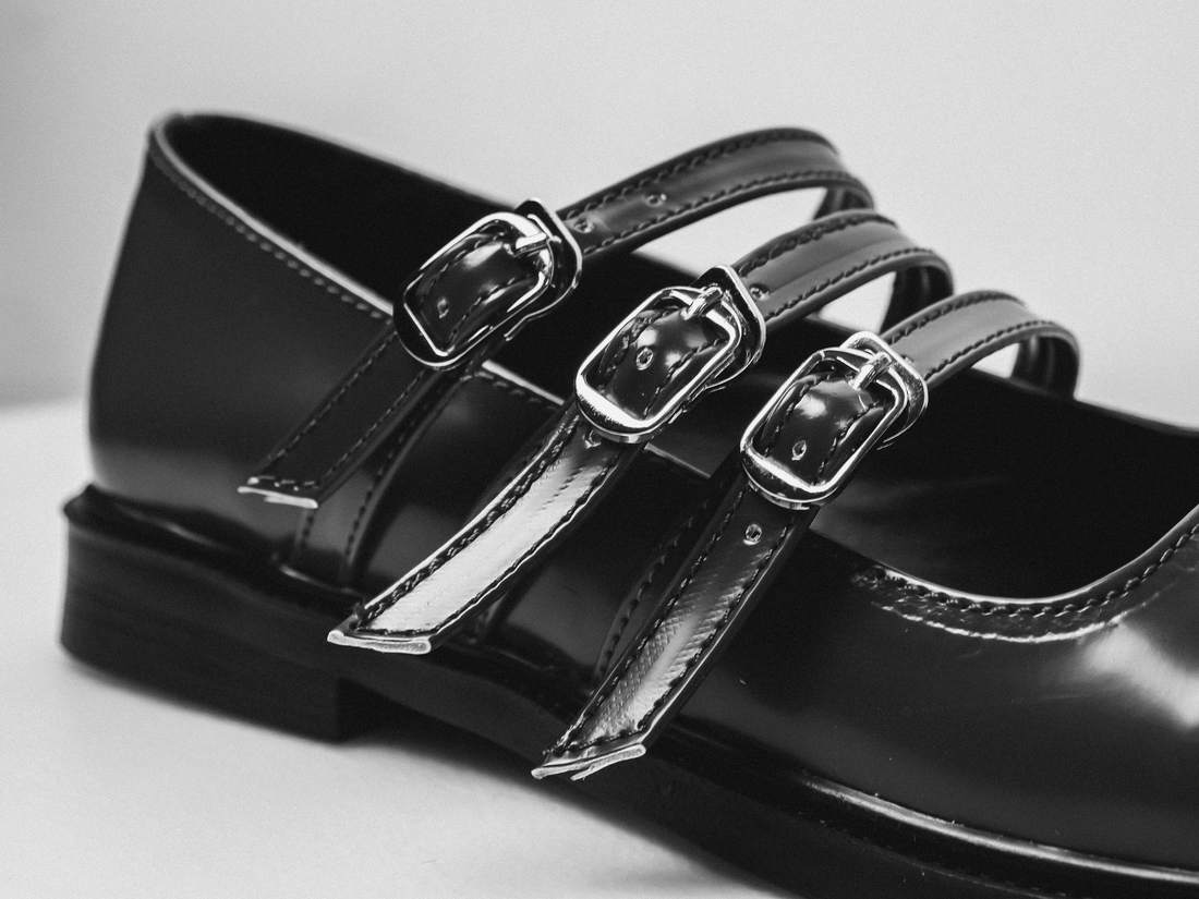 Zapato negro de charol con tres correas y hebillas plateadas, sobre un fondo claro