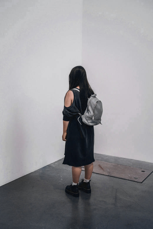 Mujer de espaldas, con vestido negro y mochila gris, de pie en una sala de muros blancos