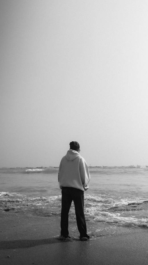 Hombre de espaldas con sudadera clara, de pie junto al mar, en blanco y negro