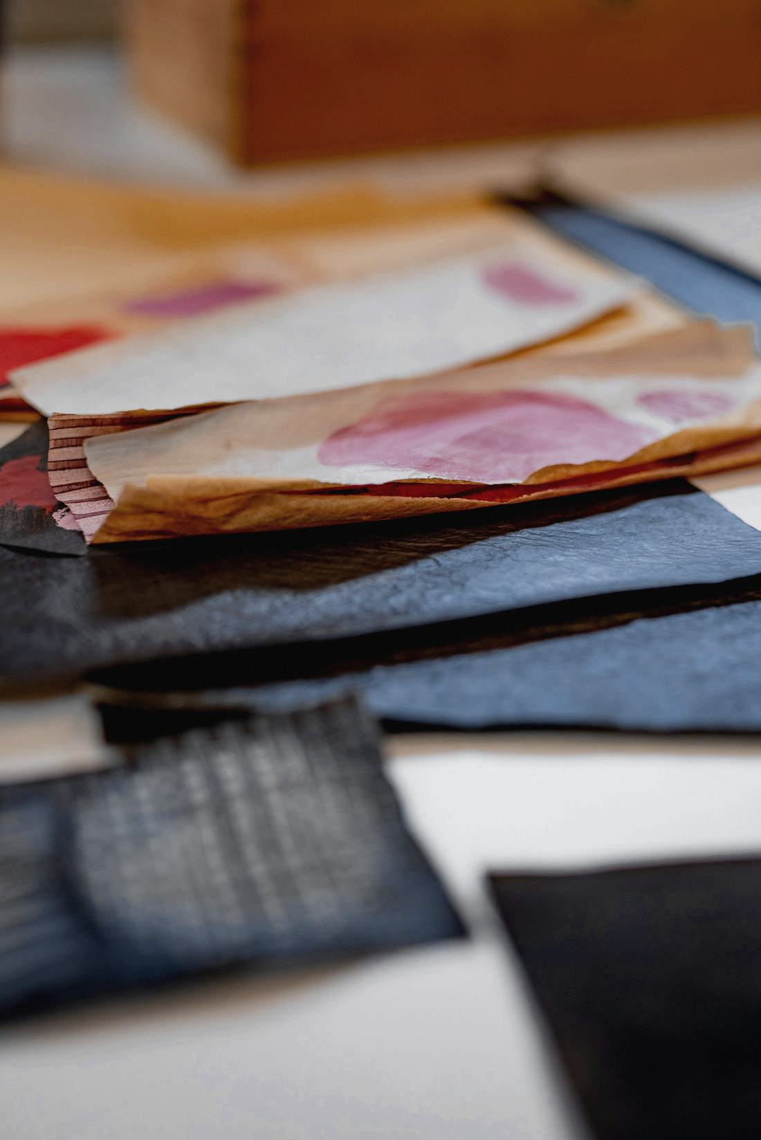 Papeles teñidos de colores y telas azules sobre una mesa, con una caja de madera desenfocada al fondo