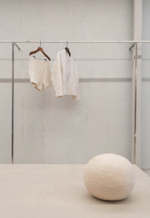 Unos shorts y una chaqueta blancos colgados de un perchero frente a un muro claro, con una esfera de fieltro sobre una mesa