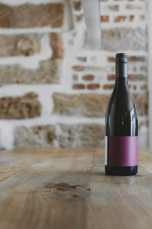 Botella de vino con etiqueta lisa sobre una mesa de madera, frente a un muro de ladrillo