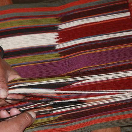 Manos tensando hilos rojos, morados y blancos sobre un telar